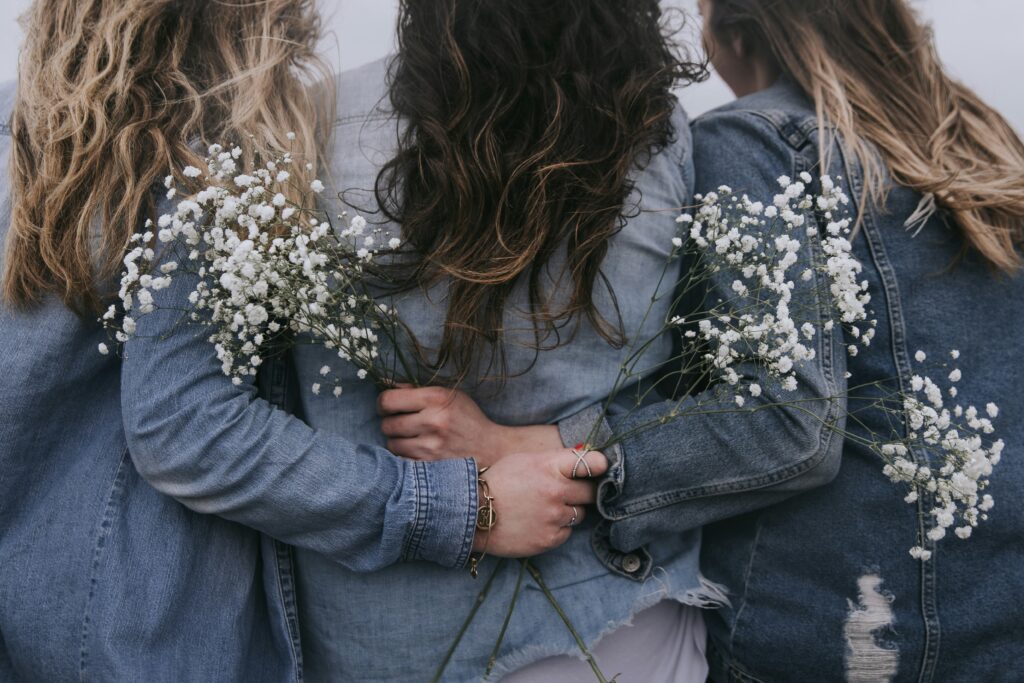 Three women standing with their back to the camera, in denim jackets and holding flowers. This is the first image on the Birthmom Buddies birth mother advocacy page.