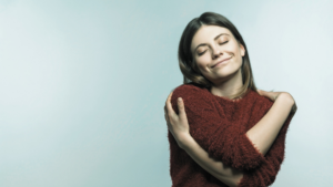 Woman embracing self-love and comfort, wearing a cozy red sweater against a light blue background.